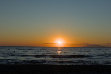 The vibrant orange sun dips towards the horizon, casting a warm glow across the sky and reflecting on the dark, rippling surface of the ocean at sunset.