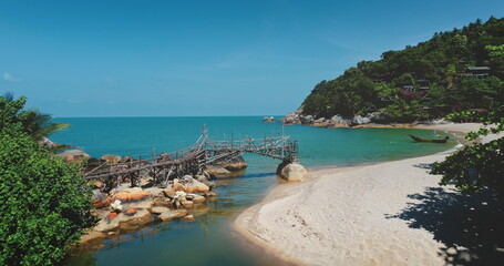 Aerial drone footage of a rickety wooden bridge connecting rocks over pristine turquoise water, leading to a serene white sand beach nestled in a lush tropical cove in Thailand