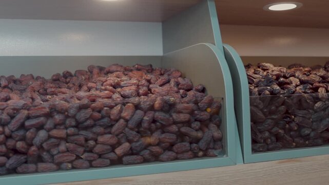 Medium to dark brown dried dates piled high in a well-lit bin with a rounded transparent cover. The fruit has a semi-gloss appearance, indicative of its moisture and freshness, and the display