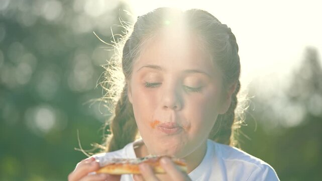 Close up of child eating pizza at picnic in park. Happy child enjoys pizza outdoors. Smiling girl savoring food during picnic. Fun park day with child eating. Child enjoying picnic meal in park.