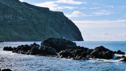 Rugged coastal rocks with a perched seagull under bright skies. A dramatic cliff rises in the...