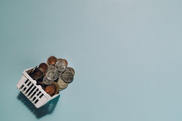 Miniature white shopping basket overflowing with coins on a calm blue background. Symbolizes saving, wealth accumulation, investment, and the value of money.
