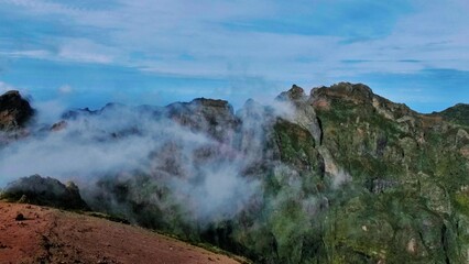 Sunlit cliffs on Madeira Island rise above misty valleys, revealing reddish rock textures and...