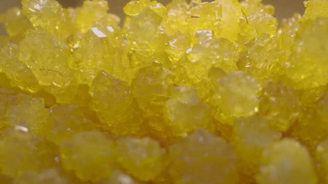 Close-up of vivid golden-yellow rock candy sticks packed together in a bin. The sugar crystals sparkle under warm lighting, emphasizing the coarse, jagged textures and rich color