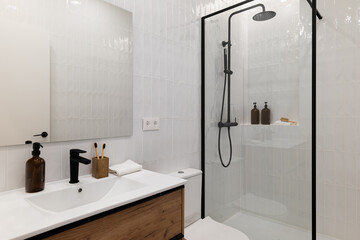 Modern bathroom with a glass shower, white tiles, wooden vanity, and black fixtures. Minimalist design with toiletries on the counter.