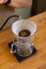 A person brews coffee using a pour-over method. The setup includes a glass dripper, coffee grounds, and a scale on a wooden table.