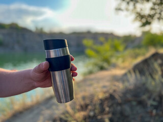 Hand holding a thermos near a lake in summer evening light