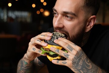 A tattooed man enjoys a tasty black burger with cheese and sesame seeds.