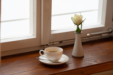 White porcelain coffee cup placed on an old wooden windowsill beside a single red rose. Cozy and...