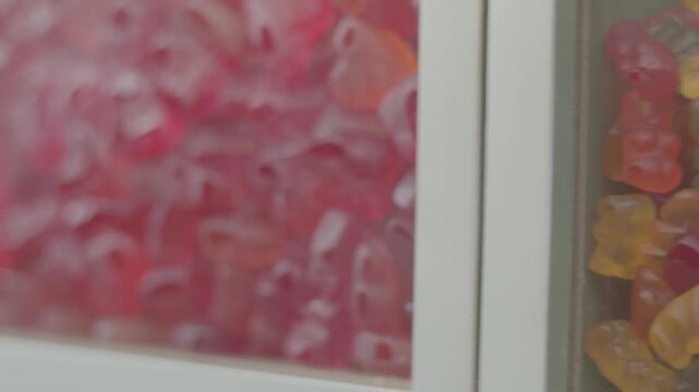 Macro shot of translucent red and orange jelly candies with a glossy surface, densely packed inside a candy bin. The candies are irregularly shaped and glow slightly under the store lighting, adding