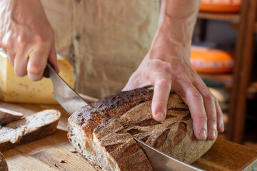 Person's hands actively slicing dark, crusty loaf of bread with long knife on wooden board, revealing lighter interior, while large piece of yellow cheese is visible in blurred background