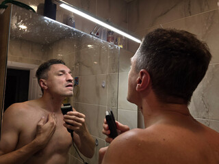 A man shaves his beard and moustache in the bathroom in front of a mirror with an electric razor