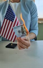 A person is holding a dollar bill that is on fire while standing beside a small American flag...
