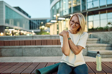 Fototapeta premium Portrait of a mature middle aged sport woman sitting with a shoulder injury in pain, after running or exercising in city park outdoors