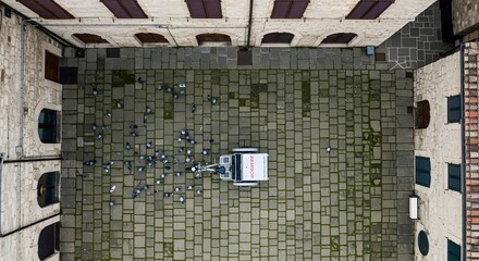 Overhead view of a generic vehicle in a stone courtyard with surrounding buildings and scattered papers creating a pattern on the ground.