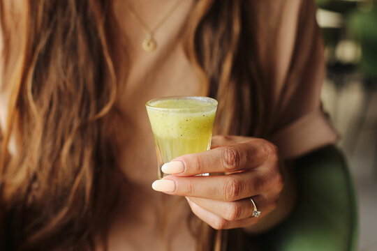 Woman holding green vitamin shot. Fresh healthy juice in small glass. Fruit smoothie in hand. Natural detox drink for energy and wellness. Blurred background. Health and beauty lifestyle detail.
