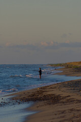 A lone individual stands in the shallow ocean water, silhouetted against a warm sunset sky, holding a fishing rod as they engage in the peaceful activity of fishing.
