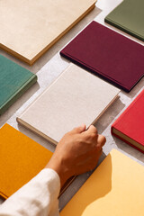 Hand picking a linen-covered book from a flat lay of colorful hardcovers on a textured background.