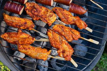 Marinated chicken fillets on wooden skewers and sausages grilling over charcoal on outdoor barbecue grill, top view.