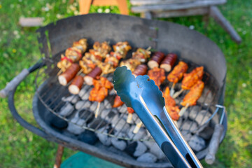 Close-up of BBQ tongs in front of a loaded charcoal grill with skewers, sausages, and marinated meat at an outdoor summer cookout.