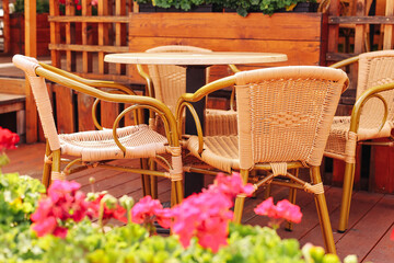 Outdoor cafe terrace with wooden table. Wicker chairs and summer flowers. Cozy restaurant setting on sunny day. Empty seats waiting for guests. Relaxing place to enjoy meal or coffee outside.