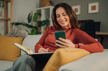 Woman taking notes from smartphone sitting on sofa at home