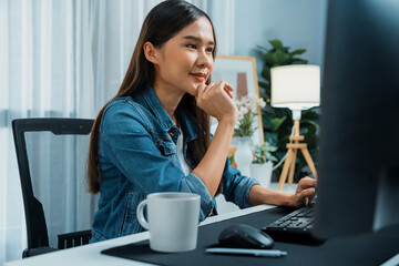Working young asian creative manager looking on pc beside hot coffee cup, creating with new website design on social media online channel planning with blue jeans shirt at home office. Stratagem.