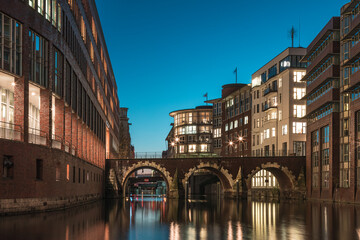 Obraz premium Blue hour long exposure of Ellerntorsbrücke spanning the Herrengrabenfleet in Hamburg, with warm lights reflecting softly on smooth water. 