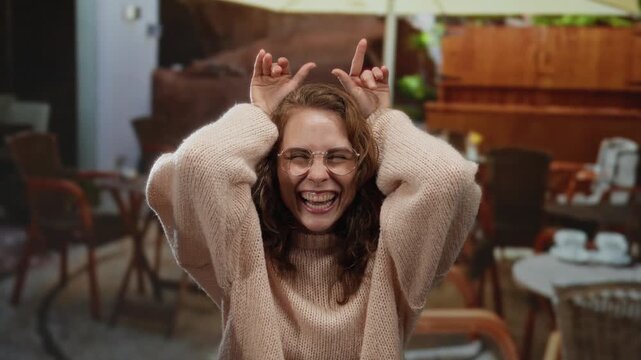 Woman making funny faces in an outdoor city cafe terrace wearing a cozy sweater and glasses showing playful silly gestures with chairs and tables in the background suggesting a relaxed vibe.