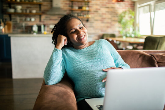 Pregnant woman relaxing on couch while video chatting on laptop at home