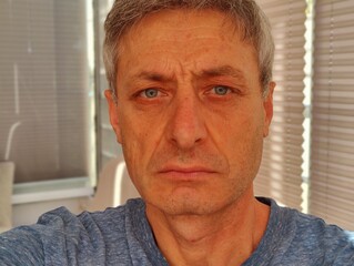 A middle-aged man with graying hair and a serious expression takes a close-up selfie in natural...