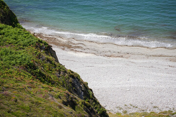 A view looking down from a green cliff onto a secluded white pebble beach with calm, clear water on the Brittany coast.