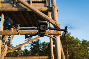 A professional camera drone with spinning propellers is hovering mid-air next to a wooden observation tower in a forest.
