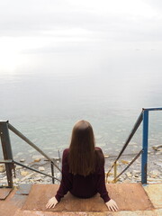 Woman sits peacefully by calm water, gazing at the horizon on an overcast morning near the shore
