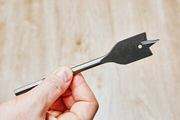 Flat drill bit with sharp cutting edges and small round hole near tip held on neutral background...