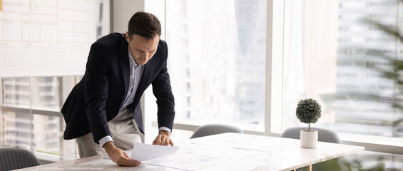 Middle-aged Hispanic businessman dressed in formal attire leaning over table, carefully examining...