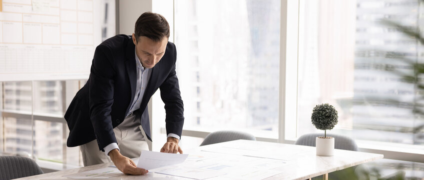 Middle-aged Hispanic businessman dressed in formal attire leaning over table, carefully examining papers with financial data spread across desk, review charts, analyzing information, horizontal image