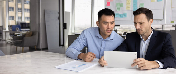 Colleagues using tablet, brainstorming ideas, collaborating on shared file or proposal, editing document or conducting research seated at conference table in modern office. Teamwork using modern tech
