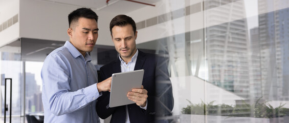 Horizontal image of two men working together using tablet in modern city highrise building office,...