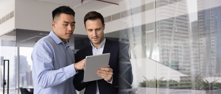 Horizontal image of two men working together using tablet in modern city highrise building office, researching, review new AI platform or application, analyzing business performance or market trends
