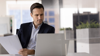 Focused man sits at desk, looks at laptop screen while holding sheet of paper, engaged in task cross-referencing digital and printed information, looks concentrated, busy in data analysis, or planning