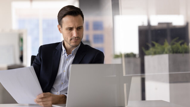 Focused man sits at desk, looks at laptop screen while holding sheet of paper, engaged in task cross-referencing digital and printed information, looks concentrated, busy in data analysis, or planning - Powered by Adobe