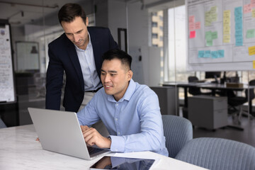 Two employees using laptop in modern workplace, discuss information displayed on screen, reviewing presentation or pitch deck, collaborating on joint, online project, analyze business data or reports