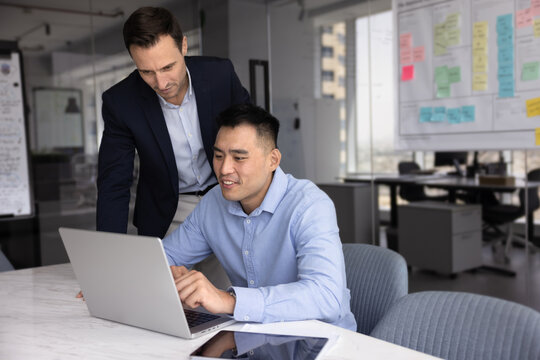 Two employees using laptop in modern workplace, discuss information displayed on screen, reviewing presentation or pitch deck, collaborating on joint, online project, analyze business data or reports
