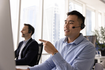 Customer service, technical support, digital communication in callcenter office. Young 30s Asian professional man wearing headset speaks while looking at computer screen, assisting to client remotely