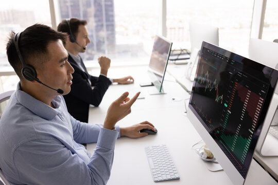 Asian professional man wear headset sits at modern desk in high-rise office, looking at monitor displaying financial trading charts involved in stock trading, financial analysis or investment advising