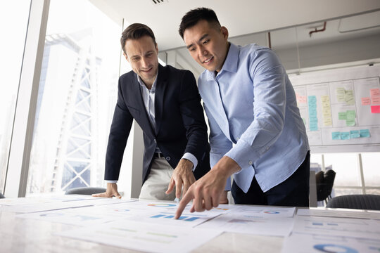 Two diverse teammates analyzing printed graphs and charts, reviewing financial performance data, discussing business metrics, collaborating on project strategy, preparing for presentation or meeting