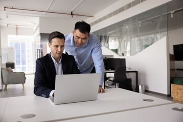 Two workmates working on joint online project using laptop, look at screen with focused expression, review financial report, analyze received data, researching information. Teamwork using modern tech