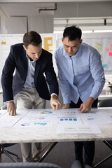 Two business professionals stand at conference table in office, deeply engaged in analyzing printed...