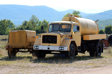 A vintage yellow water tanker truck, missing its front grille and showing visible rust and wear, stands abandoned on grass in a scenic rural field with green hills in the background